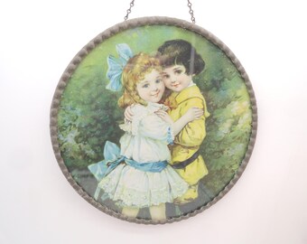 Op de afbeelding: Een ronde wandhanger met een vintage-stijl illustratie van twee kinderen die elkaar omhelzen. Het kind links draagt een witte jurk met een blauwe strik, terwijl het kind rechts een gele outfit draagt. Het object heeft een metalen frame met textuur.