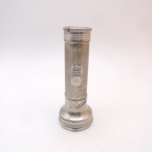 May include: A vintage silver-toned metal flashlight with a cylindrical body featuring vertical ribbed texturing. The device includes a rectangular sliding power switch on the side and a flared head section with a circular rim at the top end.