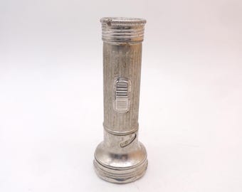 Op de afbeelding: Een vintage zaklamp van zilverkleurig metaal met een cilindervormige behuizing voorzien van verticale ribbeltextuur. Het apparaat heeft een rechthoekige schuifschakelaar aan de zijkant en een uitlopende kop met een cirkelvormige rand.