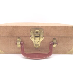 May include: A vintage tan suitcase with a textured exterior, brass-colored hardware, and a maroon handle. The corners are reinforced with gold-colored metal. The suitcase is closed with a latch.