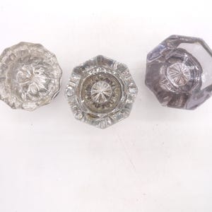 May include: Three clear glass door knobs arranged in a row. The left knob is round with a fluted edge, the center knob is octagonal with a faceted surface, and the right knob is octagonal with a smooth, slightly purple-tinted glass finish.