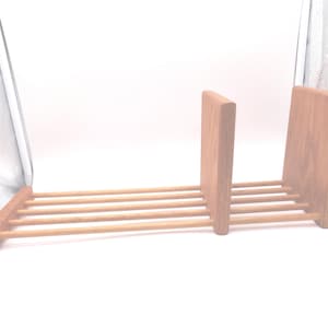 May include: A wooden book rack with a natural finish. The rack features a horizontal base with several parallel dowels and two vertical supports. The supports are connected to the base and hold books upright. The rack is designed to hold books.
