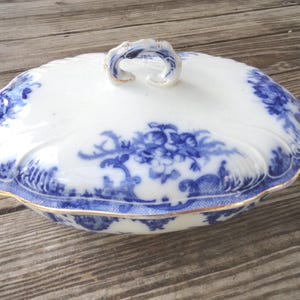May include: A white oval serving dish with a blue and white floral pattern and gold trim. The dish has a lid with a handle.