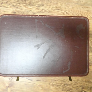 May include: A rectangular, dark brown, faux leather-covered folding tray table with rounded corners and stitched edges. The surface features visible wear and scuff marks. The table is supported by thin, metallic, gold-toned legs visible at the bottom.