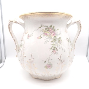 May include: A white ceramic vase with two handles, adorned with pink and yellow floral patterns and gold accents. The vase has a wide opening and a rounded body, suitable for displaying flowers or as a decorative piece.