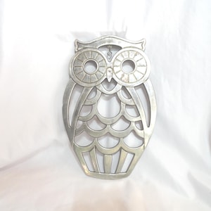 May include: Silver metal owl-shaped hot pad with a cutout design. The owl has large, round eyes with a circular pattern.
