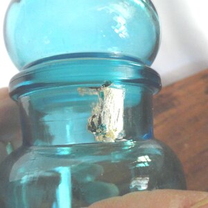May include: A vintage, teal-colored glass bottle with a rounded top and a cylindrical body. The bottle has a small, damaged label on the neck. The bottle is held by a person's hand. The bottle is likely used for storing liquids.