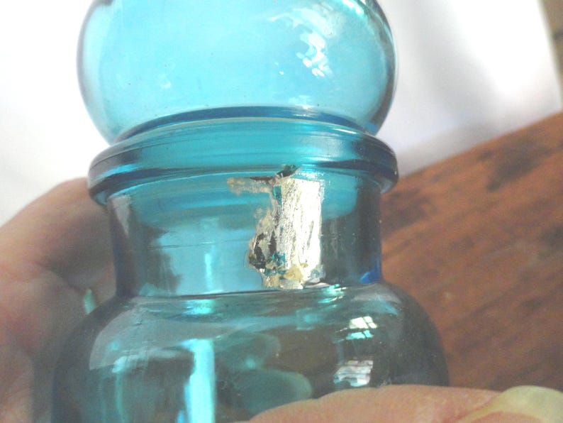 May include: A vintage, teal-colored glass bottle with a rounded top and a cylindrical body. The bottle has a small, damaged label on the neck. The bottle is held by a person's hand. The bottle is likely used for storing liquids.