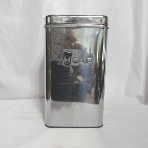 May include: A silver metal sugar canister with the word "SUGAR" embossed on the front.