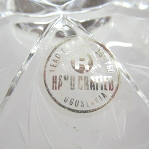 Op de afbeelding: Close-up van een helder, loodkristallen object met een ronde sticker. De sticker leest "HAND CRAFTED" en "YUGOSLAVIA" in een cirkelvormig ontwerp. Het kristal heeft een sterpatroon.