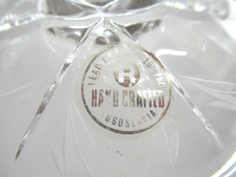 Op de afbeelding: Close-up van een helder, loodkristallen object met een ronde sticker. De sticker leest "HAND CRAFTED" en "YUGOSLAVIA" in een cirkelvormig ontwerp. Het kristal heeft een sterpatroon.