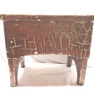 May include: A small, antique wooden stool with a rectangular top and four curved legs. The brown wood has a distressed, crackled finish with white accents, giving it a vintage appearance. The stool is likely used for seating or as a decorative piece.