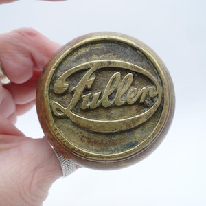 May include: A round, vintage-style door knob featuring a weathered brass finish. The center of the knob displays the word Fuller in raised, cursive lettering enclosed within an oval border. The metal surface shows signs of age, patina, and texture.