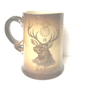 May include: A vintage ceramic beer stein with a brown handle and base. The stein features a detailed illustration of a stag with antlers, the number 19, and the text "A.C. Dennis". The background has a gradient of brown and tan.