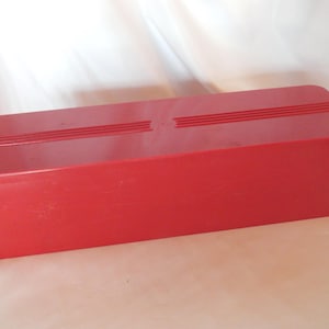 May include: A red plastic storage box with a smooth, flat top and a slightly textured bottom. The box has a rectangular shape and is approximately 12 inches long.
