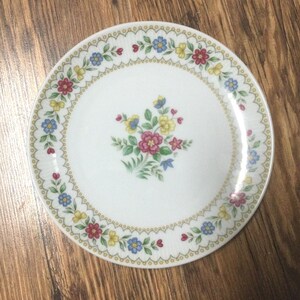 May include: White ceramic plate with a floral design. The plate features a central bouquet of pink, yellow, and blue flowers, surrounded by a border of smaller flowers and a gold trim. The plate is round.