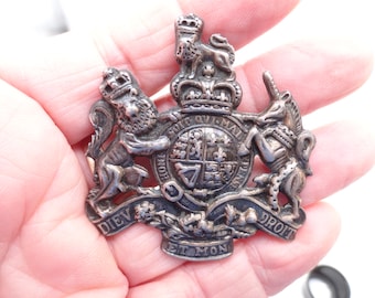 Op de afbeelding: Een zilverkleurige metalen badge met het koninklijk wapen van het Verenigd Koninkrijk. Het ontwerp bevat een leeuw en een eenhoorn die een centraal schild geflankeerd door een kroon omringen, met het motto DIEU ET MON DROIT en HONI SOIT QUI MAL Y PENSE.