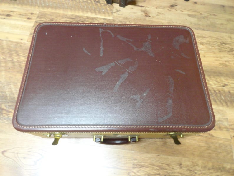 May include: A rectangular suitcase featuring a dark brown textured top lid with visible surface wear and scuff marks. The base of the suitcase is a contrasting light tan color, accented with brass-toned metal latches and a dark brown handle.