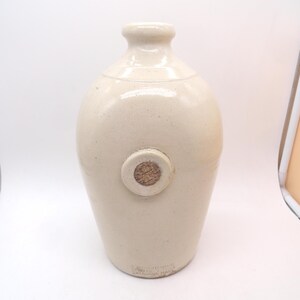 May include: A cream-colored ceramic jug with a smooth, glossy finish and a narrow neck. The body features a circular indentation containing a textured cork plug. Near the base, there is faint, embossed text stamped into the ceramic material.