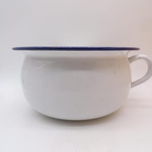 May include: A white enamel chamber pot with a rounded body and a single side handle. The rim of the pot is finished with a dark blue border. The white surface features subtle, scattered brown speckles throughout the exterior of the vessel.