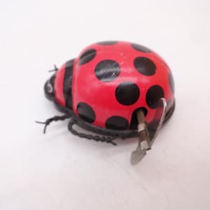 May include: A wind-up toy shaped like a ladybug with a bright red domed shell featuring black circular spots. The toy has a black head, thin black wire legs, and a silver metal winding key protruding from the side of its rounded body.