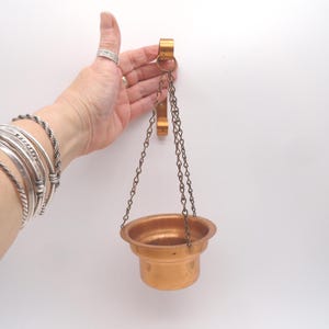 May include: A copper-colored hanging incense burner with a round bowl shape, suspended by chains from a wall-mounted bracket. The burner is designed to hold incense and is made of metal. The chains and bracket are also copper-colored.