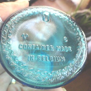 May include: A close-up of the bottom of a teal-colored glass container. The text "CONTAINER MADE IN BELGIUM" is embossed on the glass. The container has a textured, decorative edge.