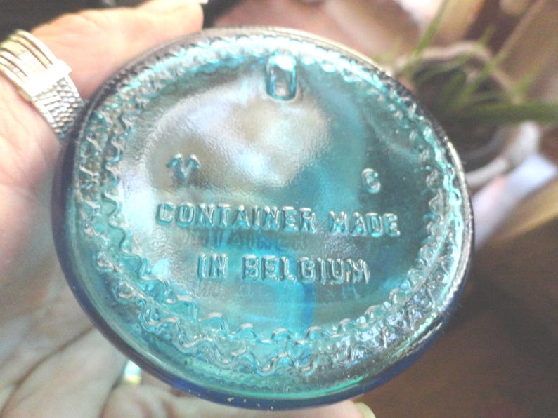 May include: A close-up of the bottom of a teal-colored glass container. The text "CONTAINER MADE IN BELGIUM" is embossed on the glass. The container has a textured, decorative edge.