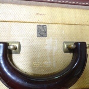 May include: A vintage suitcase with a textured cream-colored exterior, featuring a dark brown curved handle attached with brass-toned hardware. A small rectangular metal emblem displays the text U.S. TRUNK CO. and the letters S.G. are embossed below.