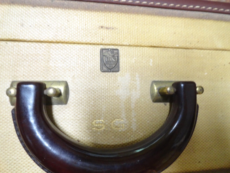 May include: A vintage suitcase with a textured cream-colored exterior, featuring a dark brown curved handle attached with brass-toned hardware. A small rectangular metal emblem displays the text U.S. TRUNK CO. and the letters S.G. are embossed below.