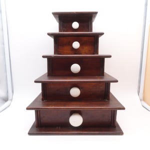 May include: A dark brown wooden tiered storage unit featuring five stacked drawers of decreasing size from bottom to top. Each drawer is equipped with a single round white knob centered on the front panel, creating a pyramid-like structure.