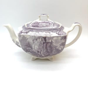 May include: A vintage purple and white ceramic teapot with a lid. The teapot features a detailed landscape scene and rests on three small feet. The spout and handle are white, and the overall design is classic and elegant.