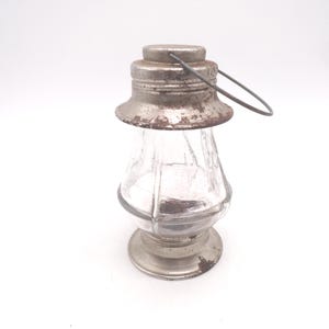 Pu&ograve; includere: Una lanterna in vetro vintage con coperchio e base in metallo. Il corpo in vetro trasparente ha un design sfaccettato. I componenti metallici mostrano segni di et&agrave; e usura, con una maniglia.