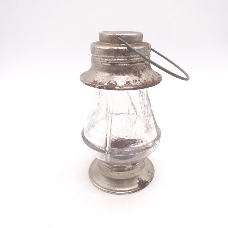 Pu&ograve; includere: Una lanterna in vetro vintage con coperchio e base in metallo. Il corpo in vetro trasparente ha un design sfaccettato. I componenti metallici mostrano segni di et&agrave; e usura, con una maniglia.