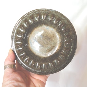 May include: A handmade, ceramic bowl with a dark brown exterior and a lighter interior. The rim features a repeating, textured pattern. The bowl is held in a hand, showcasing its size and shape. The glaze has a glossy finish.