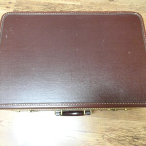 May include: A rectangular, dark brown hard-shell suitcase with rounded corners and visible stitching along the edges. The suitcase features a dark brown handle and metallic gold-toned hardware latches positioned along the front side.