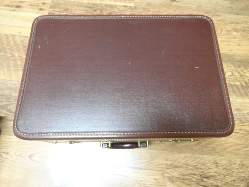 May include: A rectangular, dark brown hard-shell suitcase with rounded corners and visible stitching along the edges. The suitcase features a dark brown handle and metallic gold-toned hardware latches positioned along the front side.