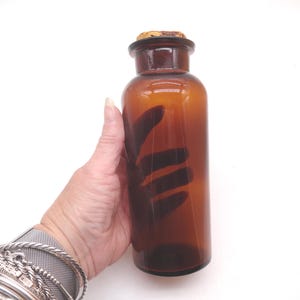 May include: A cylindrical amber glass bottle with a smooth, translucent finish and a wide neck. The bottle is topped with a natural, light brown cork stopper that fits securely into the opening. The glass is clear and uniform in color.