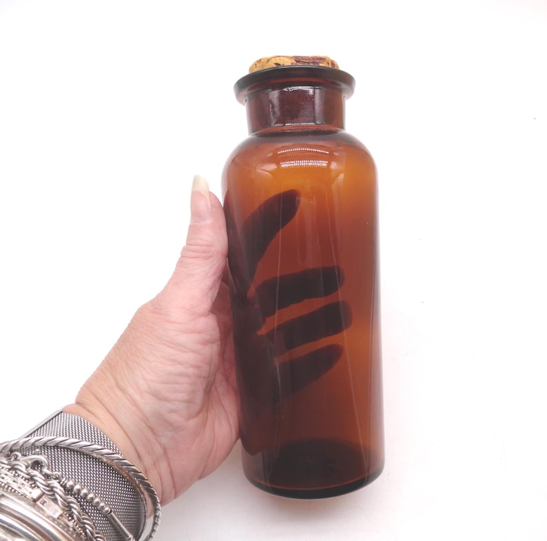 May include: A cylindrical amber glass bottle with a smooth, translucent finish and a wide neck. The bottle is topped with a natural, light brown cork stopper that fits securely into the opening. The glass is clear and uniform in color.