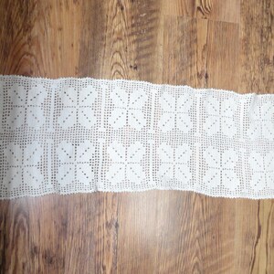 May include: A rectangular white crochet table runner featuring a repeating pattern of square motifs. Each square contains a four-petaled floral design created with openwork stitches, connected by a grid-like mesh border throughout the piece.