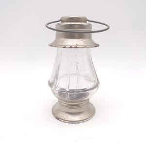 May include: A small, vintage-style glass lantern with a silver-colored metal base, top, and handle. The clear glass body has a rounded shape with vertical ridges. The lantern is designed to hold a small candle or light source.