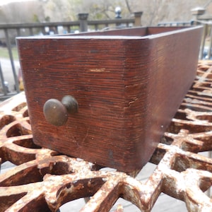 May include: A wooden drawer with a brass knob. The drawer is made of dark wood with a grain pattern. The drawer is sitting on a metal surface.