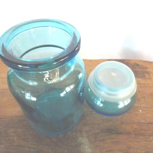 May include: A clear, teal-colored glass jar with a rounded body and a wide, flared rim. Beside it is a matching, smaller jar with a lid. The jars are sitting on a wooden surface.