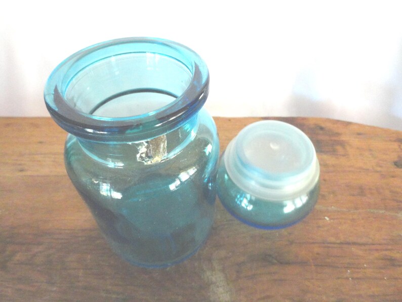 May include: A clear, teal-colored glass jar with a rounded body and a wide, flared rim. Beside it is a matching, smaller jar with a lid. The jars are sitting on a wooden surface.