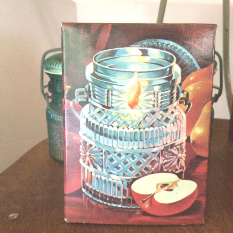 May include: A vintage glass candle jar with a decorative diamond pattern. The jar is filled with a white candle and has a flame burning inside. The jar is surrounded by fruit, including a red apple and a pear.