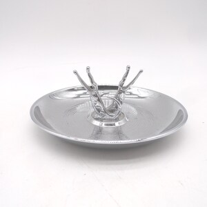 May include: A silver-colored serving dish with a decorative, abstract figure in the center. The dish has a shallow, round shape with a textured surface. The figure appears to be a stylized representation of a person or animal.