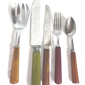 May include: A set of vintage silverware including a serving fork, a butter knife, a knife, a fork, and a spoon. The handles are in shades of brown, green, and burgundy. The metal parts are silver.