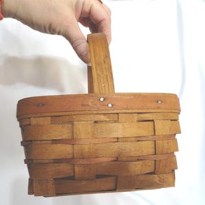 May include: A small, brown wooden basket with a woven design and a single handle. The basket has a light brown rim.