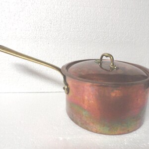 May include: A copper saucepan with a brass handle and lid. The pot has a rounded body and a long, slender handle. The lid has a small brass handle. The copper has a reddish-brown color with some green and gold hues.