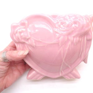 May include: A glossy light pink ceramic planter shaped like a heart, featuring raised relief details of roses and leaves on the front surface. The vessel is supported by two small, integrated feet at the base, creating a stable, elevated structure.